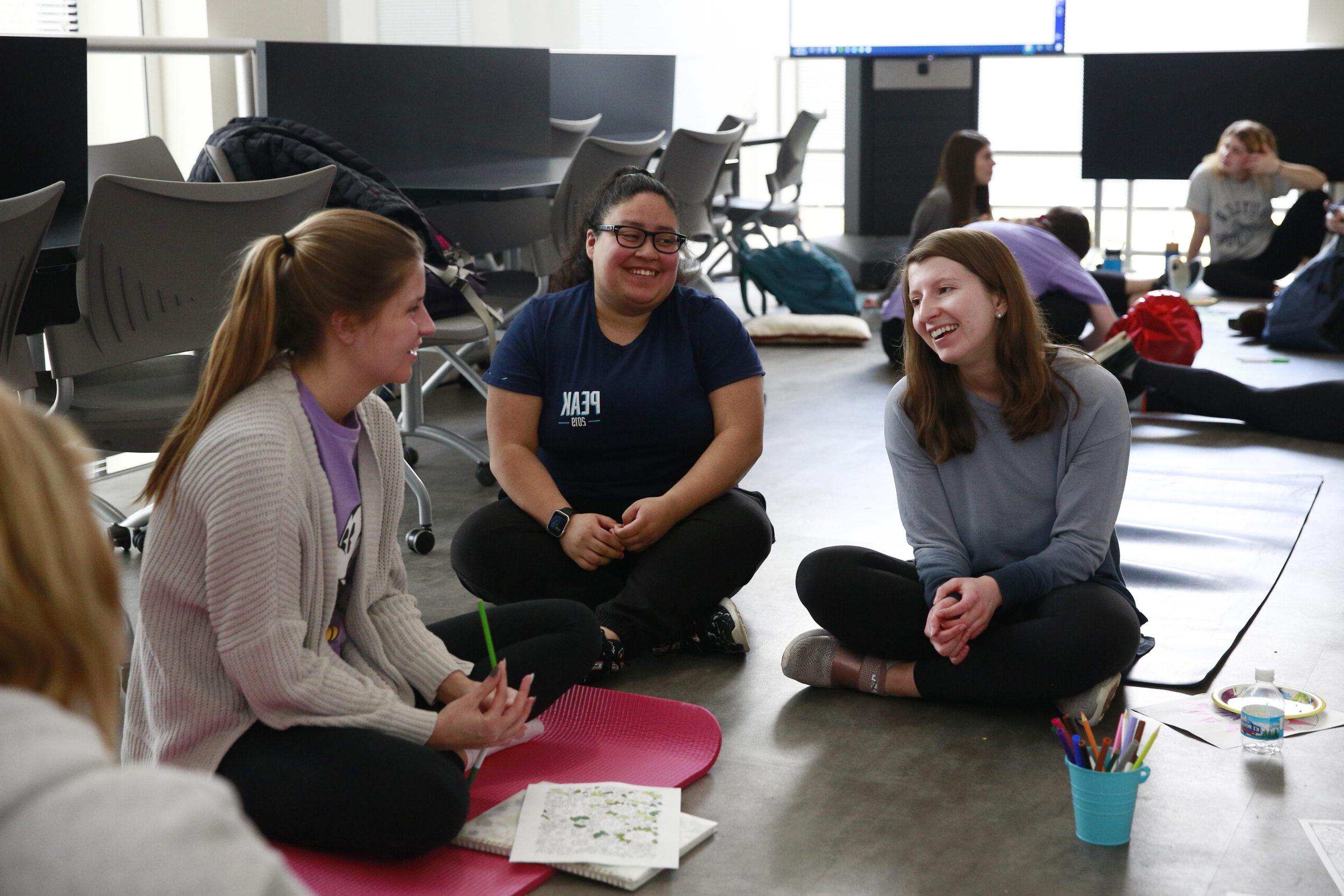 Three female student sit in a circle on the floor and are working on a project.